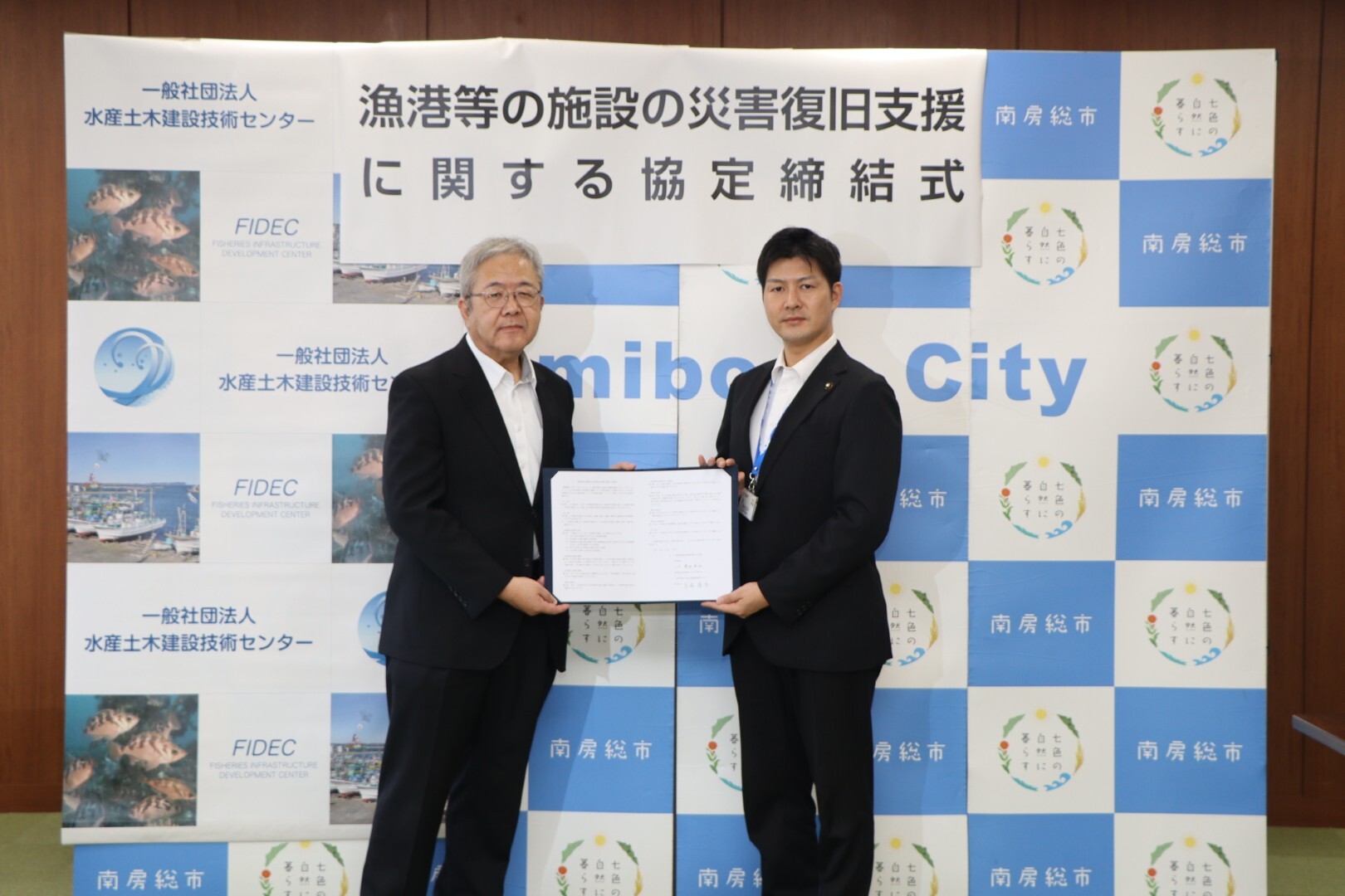 スーツ姿の二人の人物が協定書を掲げて市のロゴと災害復旧支援に関する横断幕の前で並んでいる